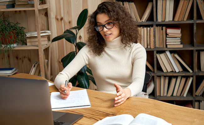 a young woman sitting in front of a laptop taking notes in a notebook